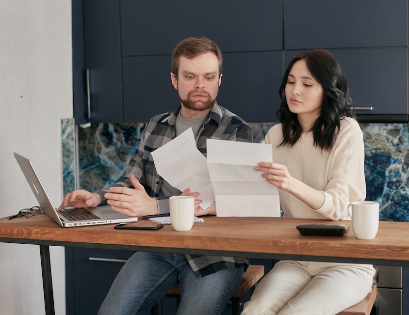 A couple sitting at a wooden table, reviewing documents with a laptop nearby, expressing concern.